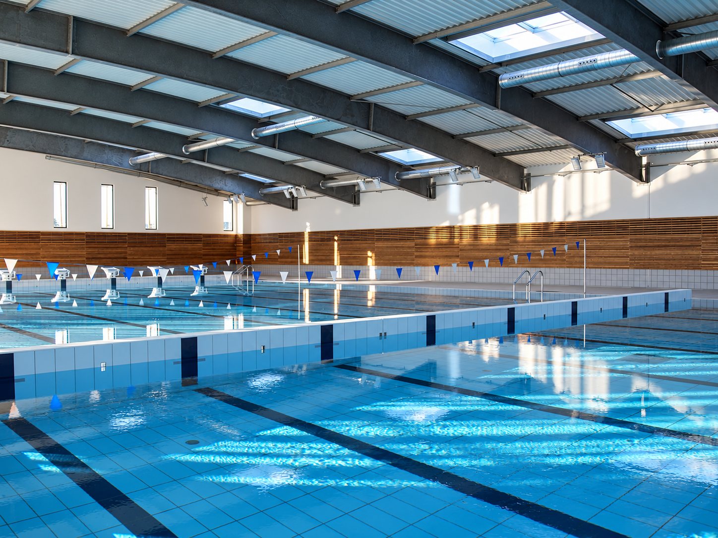 Intérieur d'une piscine couverte lumineuse avec bassin carrelé, plots de départ, fanions bleus et blancs et charpente métallique voûtée.