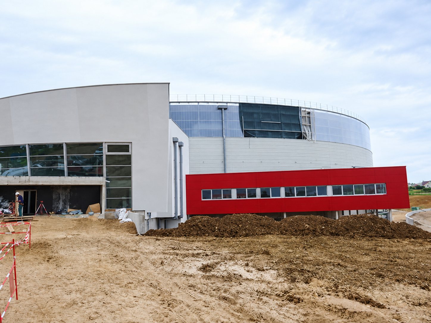 Chantier d'un grand bâtiment arrondi à façade grise et bandeau rouge, entouré de terre remuée et de barrières de chantier.