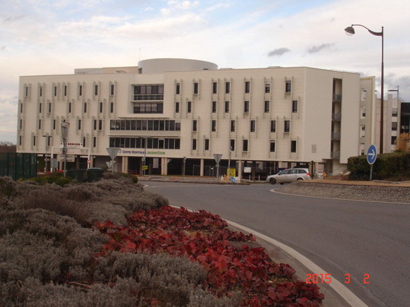 Un grand bâtiment blanc à plusieurs étages donne sur un rond-point planté de lavande et de feuillage rouge, avec une voiture garée devant.