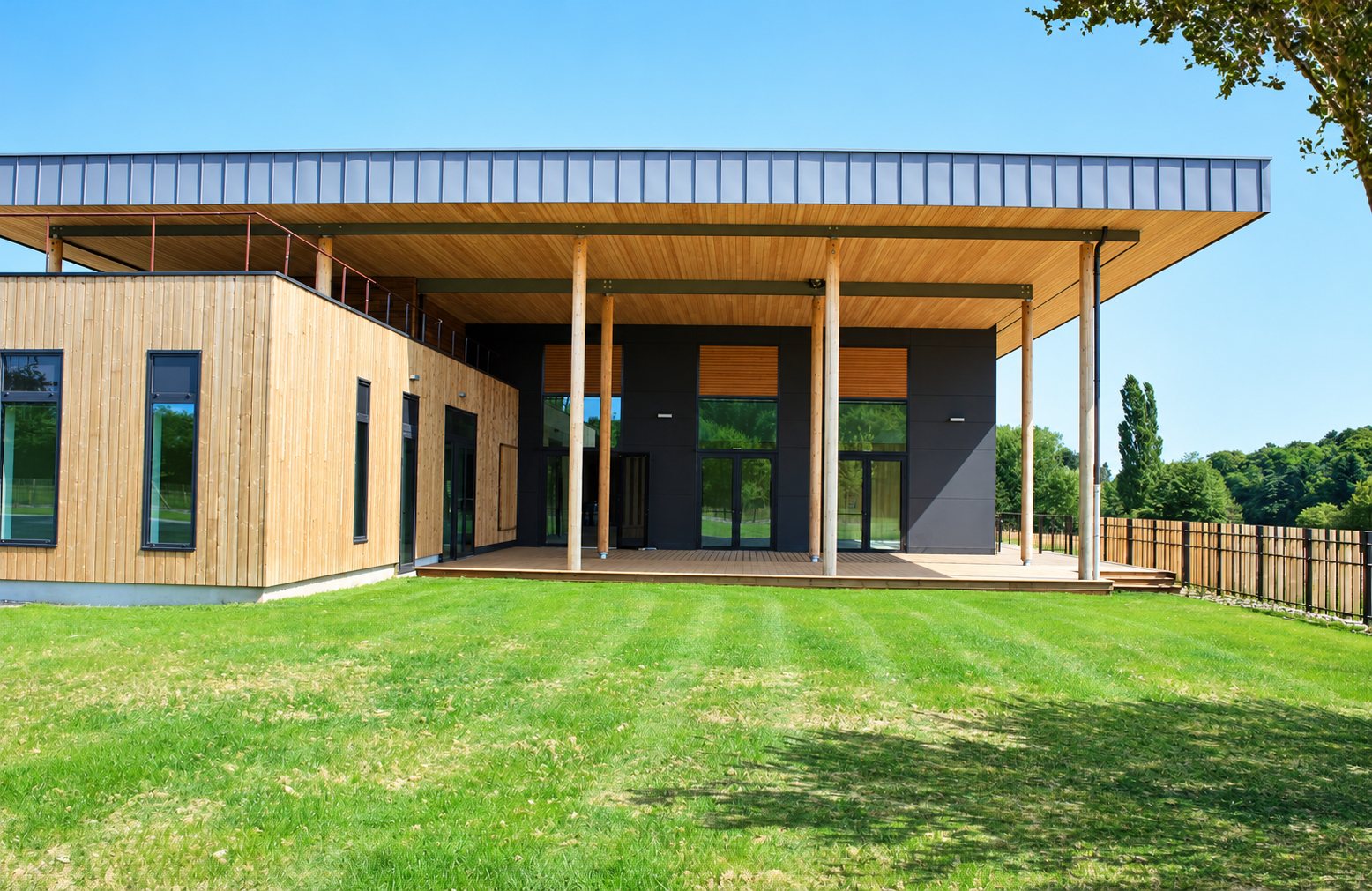 Bâtiment contemporain à bardage en bois et panneaux sombres, avec un large toit en débord soutenu par de fins poteaux, entouré d'une pelouse et d'une clôture en bois.