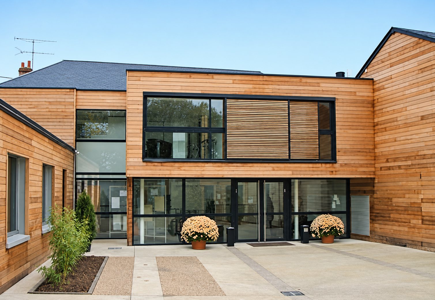 Façade d'un bâtiment contemporain revêtu de bardage en bois clair, avec de grandes baies vitrées et deux pots de chrysanthèmes dans la cour dallée.