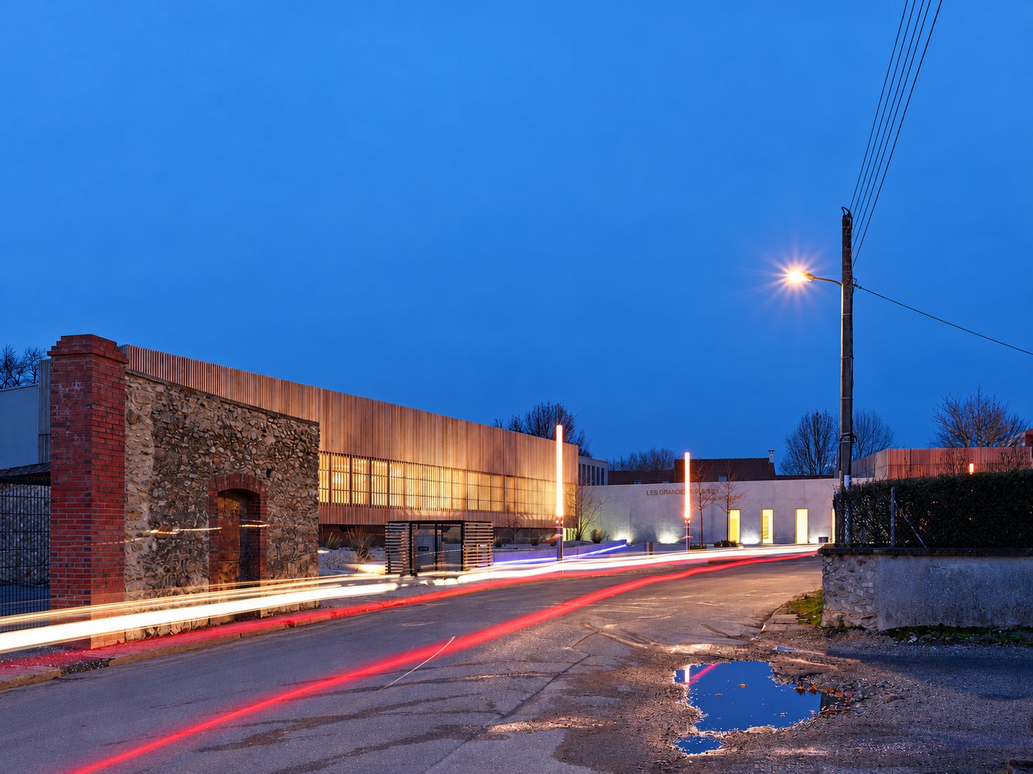 Au crépuscule, un bâtiment contemporain à façade de bois éclairée longe une route, à côté d'un vieux mur de pierre et de brique, avec des traînées lumineuses de voitures.