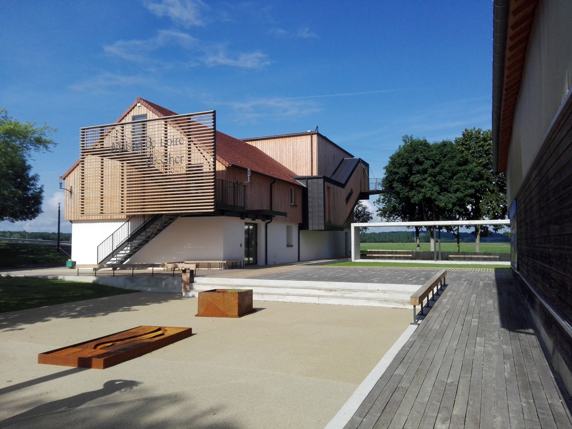 Un bâtiment contemporain à façade en bois et toit de tuiles rouges, avec un escalier métallique, donne sur une esplanade et une terrasse en bois sous un ciel bleu.