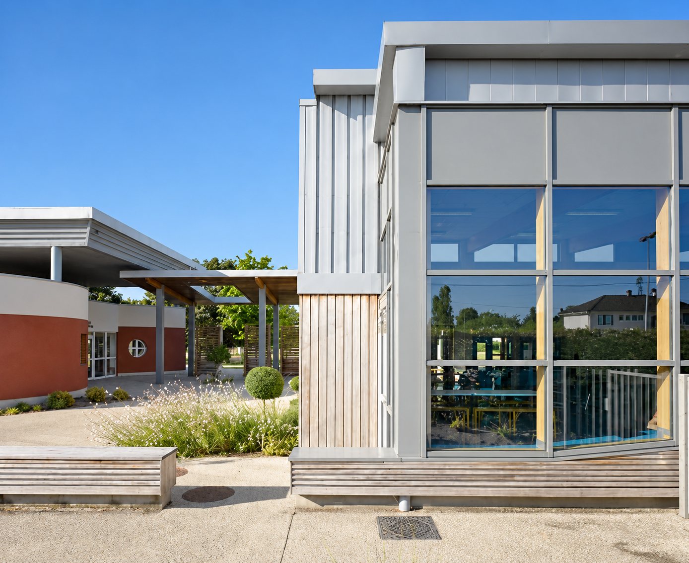 Façade d'un bâtiment contemporain à bardage bois et métal gris, avec de grandes vitres, un auvent et un jardin planté au premier plan.