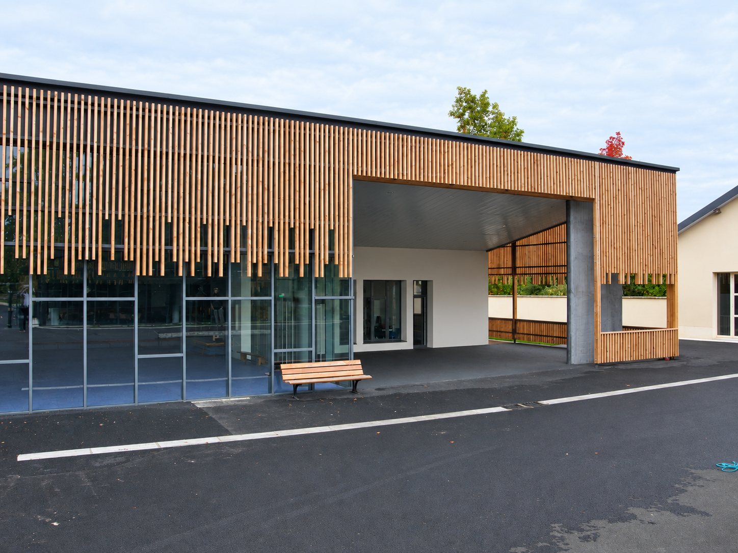 Bâtiment contemporain à façade vitrée habillée de lames de bois verticales, avec un préau couvert et un banc en bois devant.