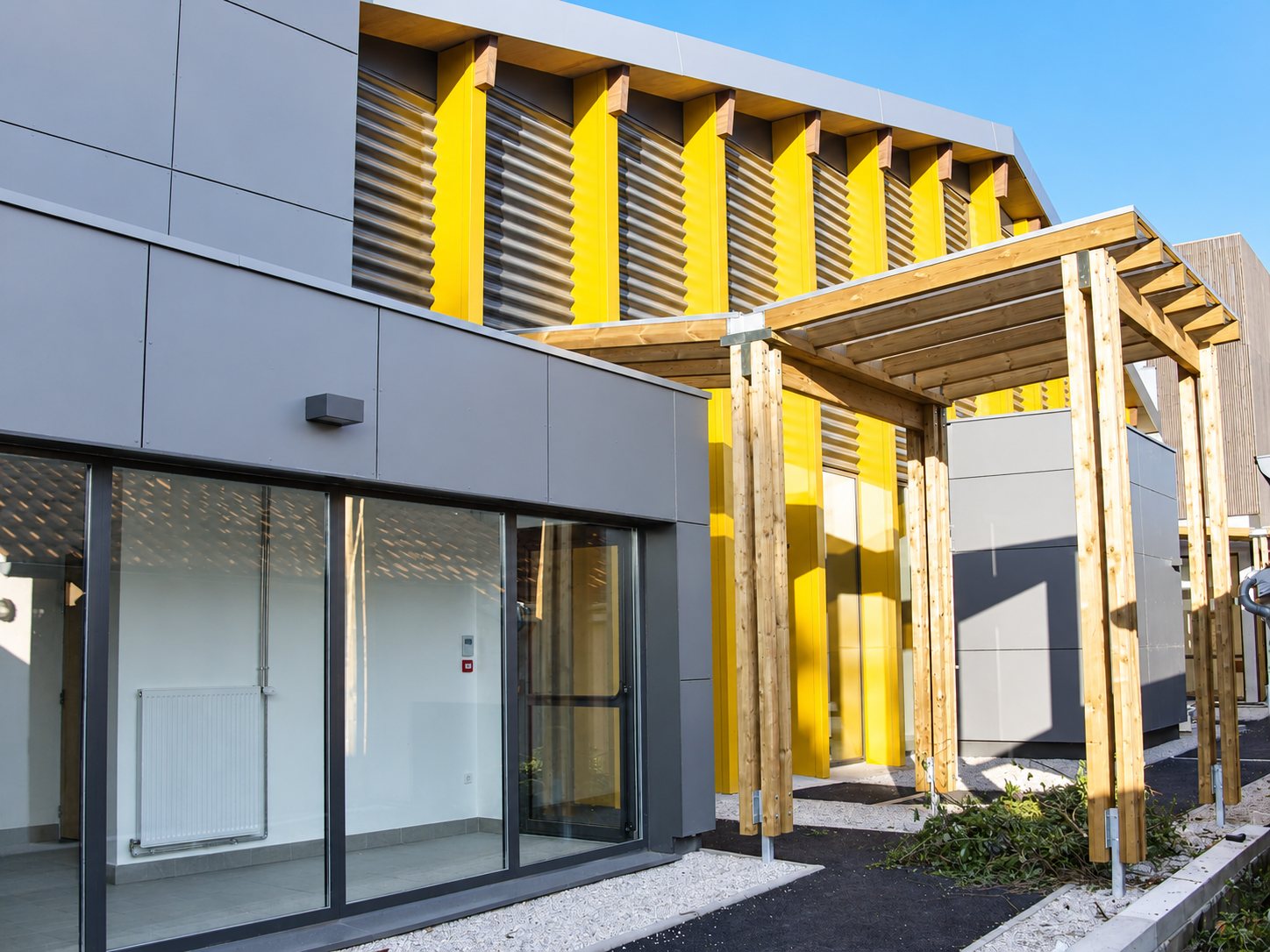 Façade d'un bâtiment récent aux panneaux gris et aux montants jaunes, avec grandes baies vitrées et pergola en bois au premier plan.