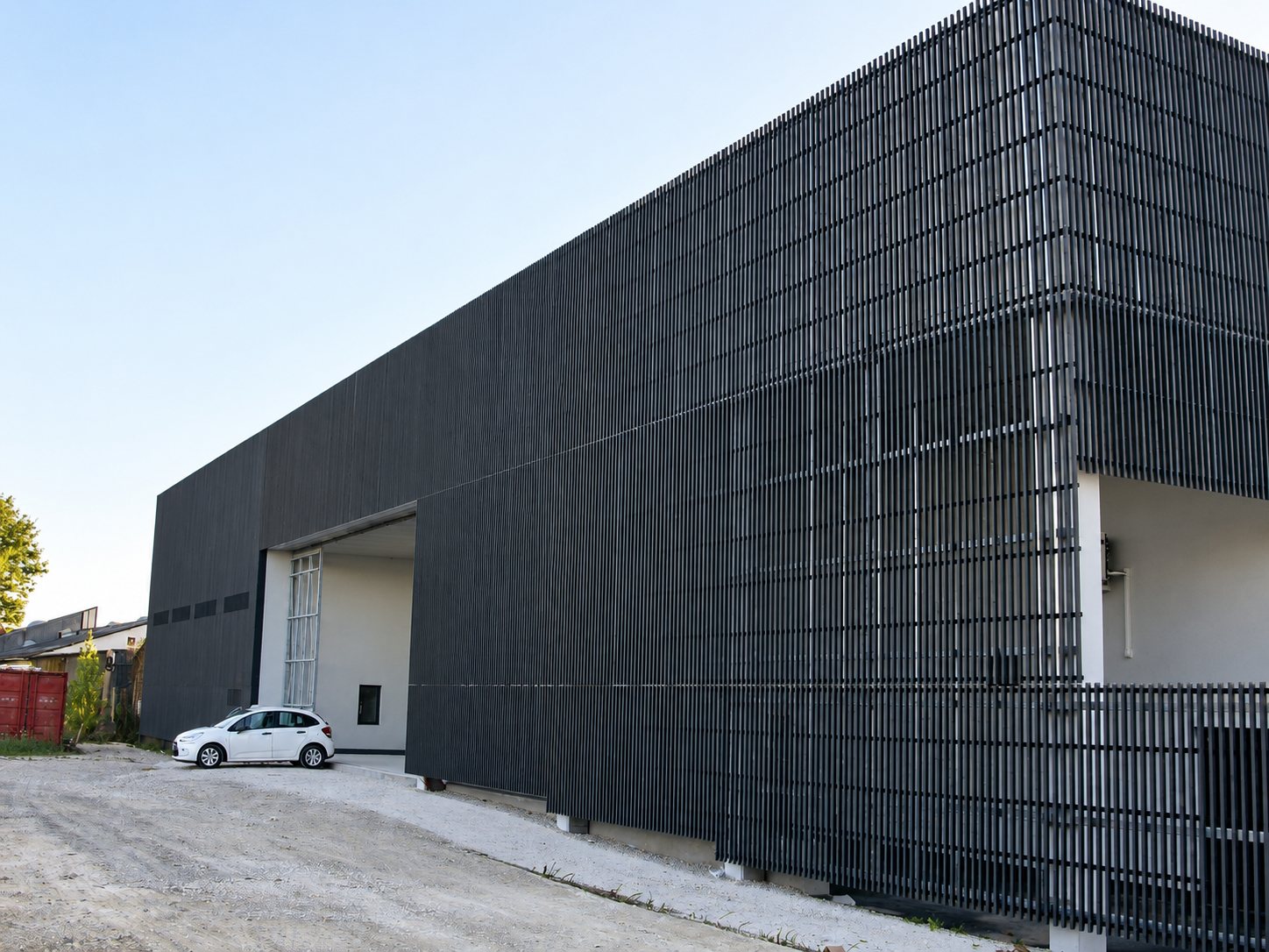 Grand bâtiment moderne recouvert de lames verticales sombres, avec une voiture blanche garée devant l'entrée sur un chemin de gravier.