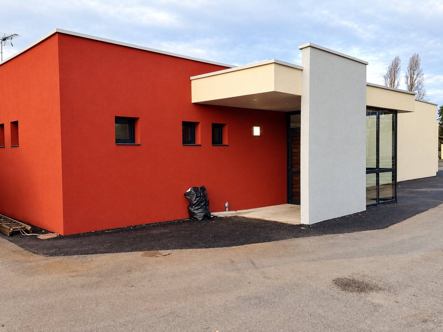 Construction récente de plain-pied aux murs rouge brique et crème, avec grandes baies vitrées et sol en enrobé noir fraîchement posé.
