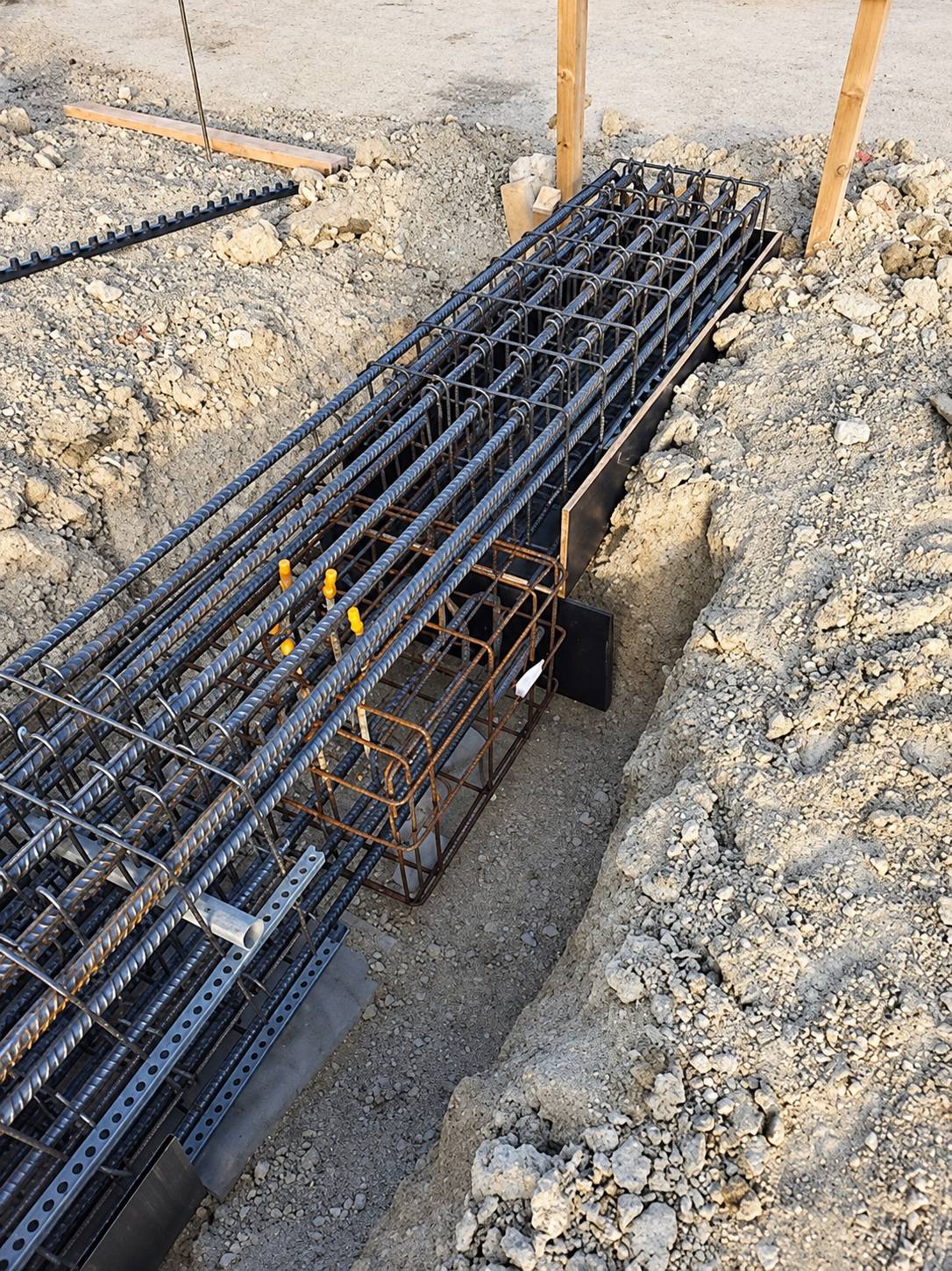 Des cages d'armature en acier sont posées dans une tranchée creusée dans un sol rocailleux, sur un chantier de construction.