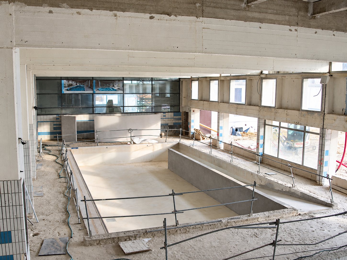 Vue en hauteur d'un bassin de piscine intérieure vide et en travaux, entouré de garde-corps et de grandes baies vitrées.