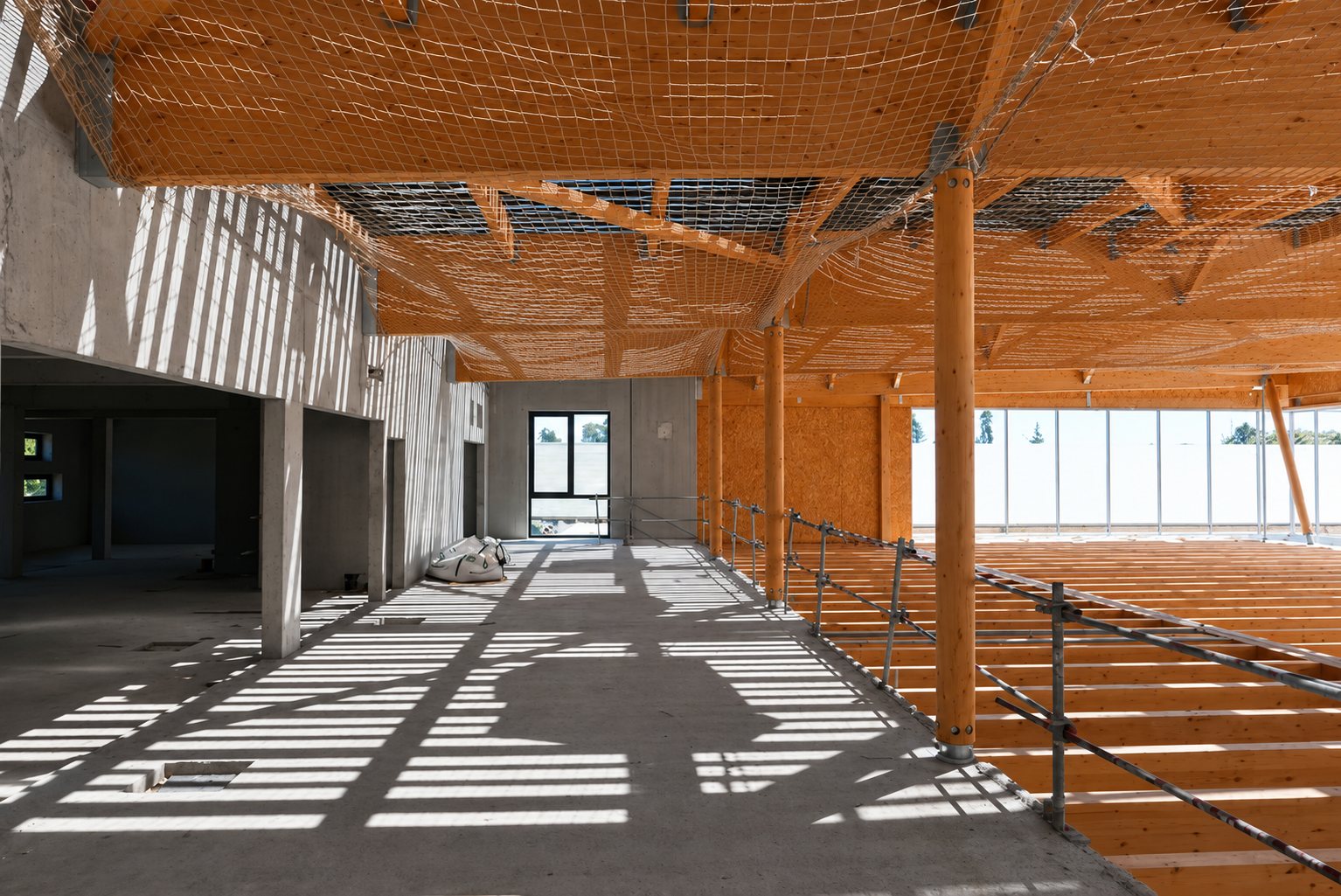 Intérieur d'un bâtiment en construction avec charpente et poteaux en bois, filet de sécurité tendu au plafond et lumière projetant des ombres rayées sur la dalle en béton.