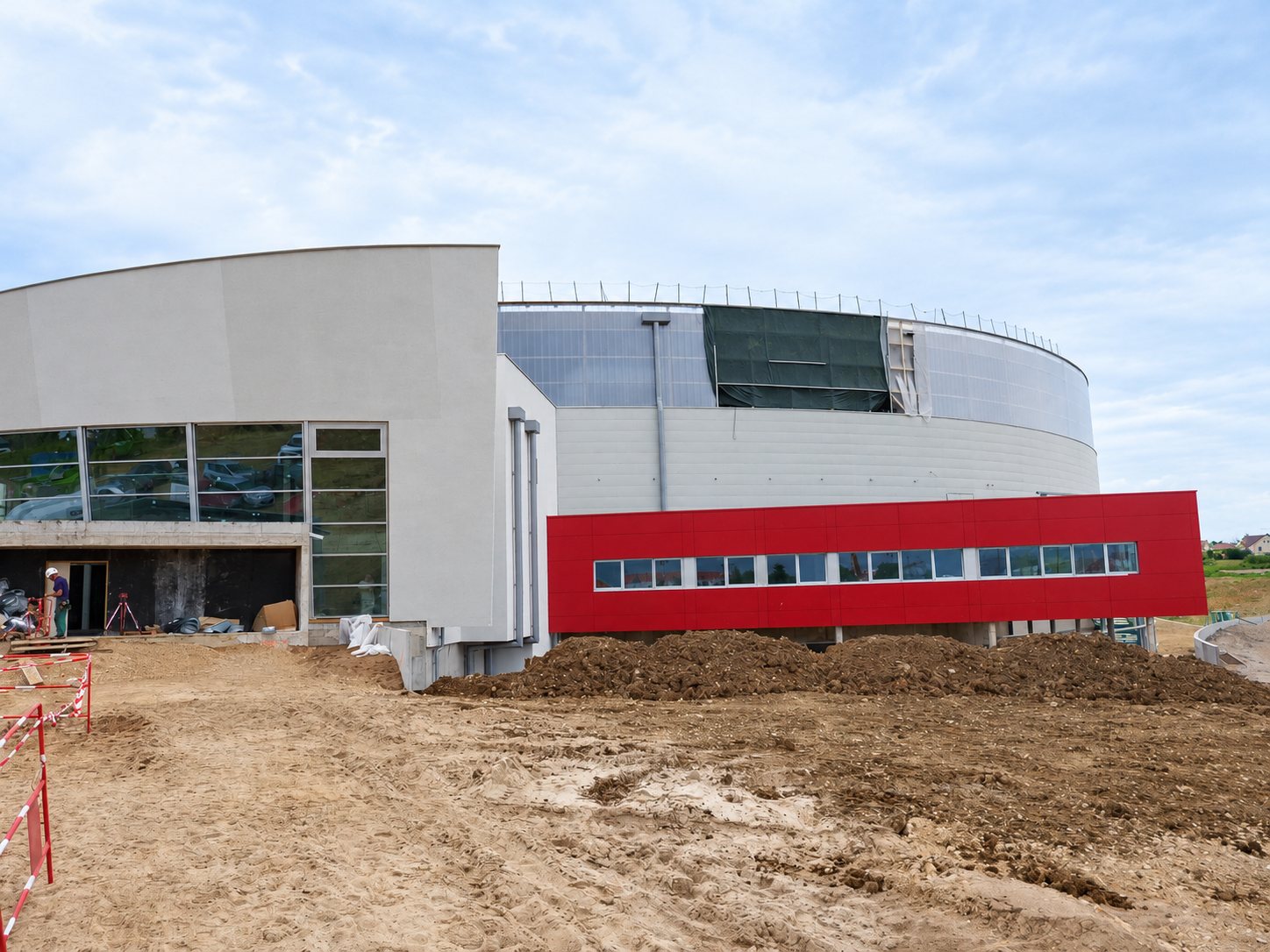 Un grand bâtiment circulaire en construction, avec des façades grises et rouges, entouré de terre remuée et de barrières de chantier rouges et blanches.
