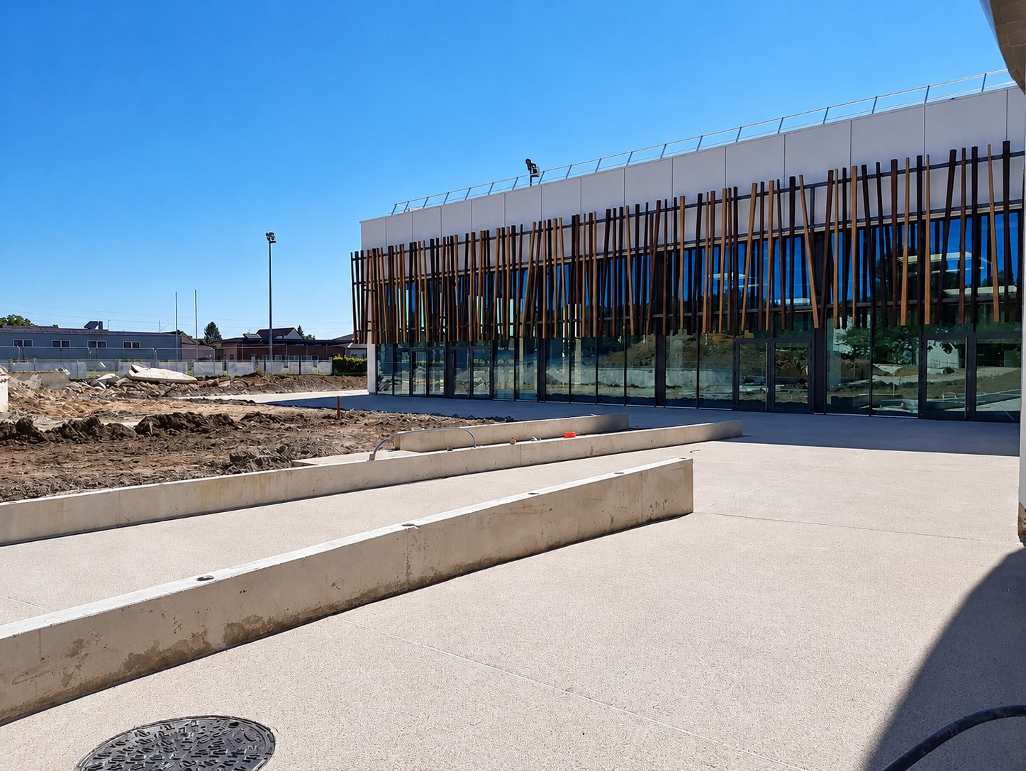 Un bâtiment moderne à façade vitrée ornée de lames de bois, devant un parvis en béton clair et des terrassements en cours sous un ciel bleu.