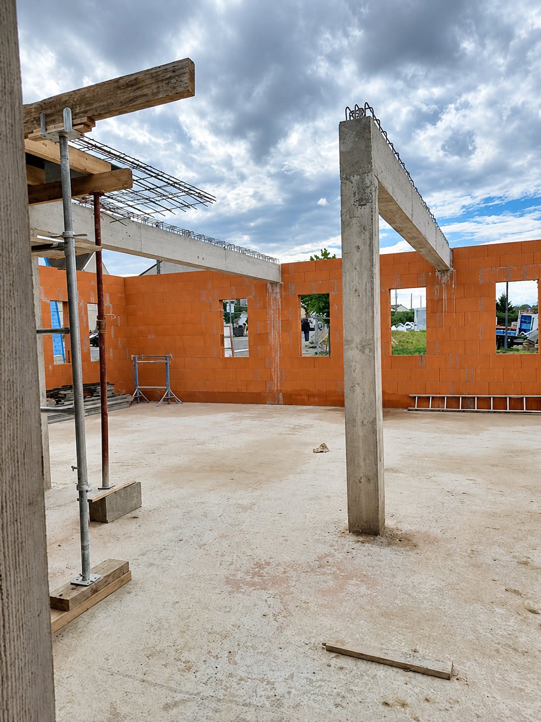 Chantier d'une maison en construction avec des murs en briques rouges, des poteaux et poutres en béton, et une dalle brute sous un ciel nuageux.