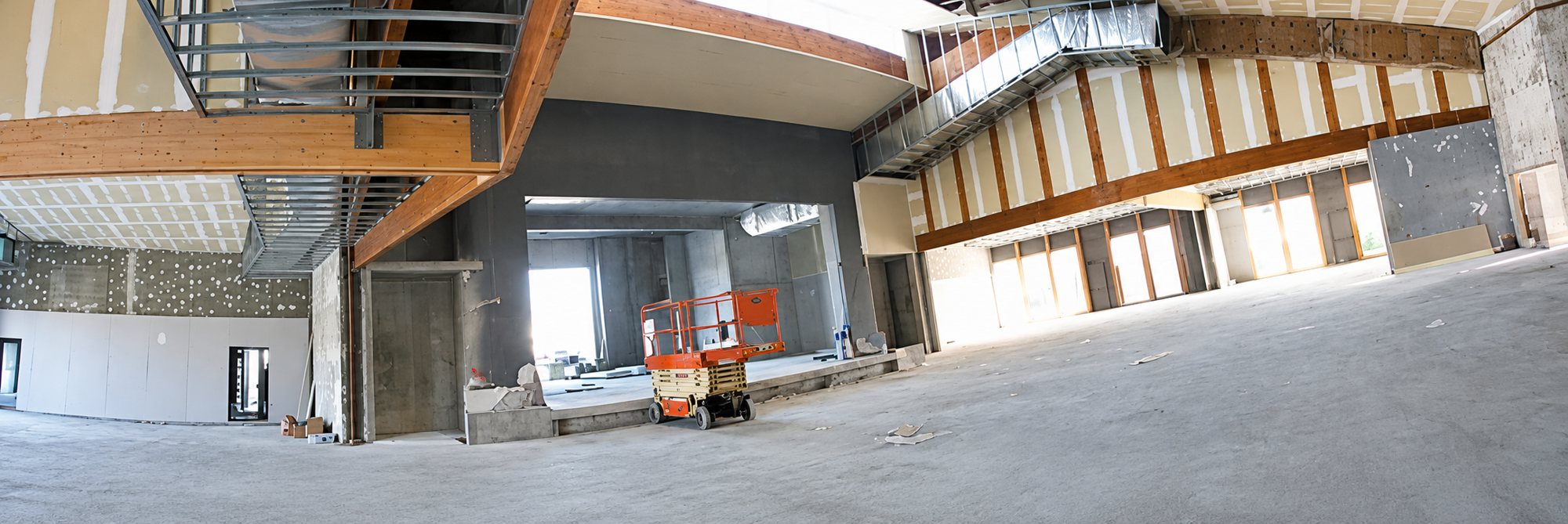 Vue panoramique d'un grand volume intérieur en travaux, avec des murs en plaques de plâtre, des structures en bois et une nacelle élévatrice orange au centre.
