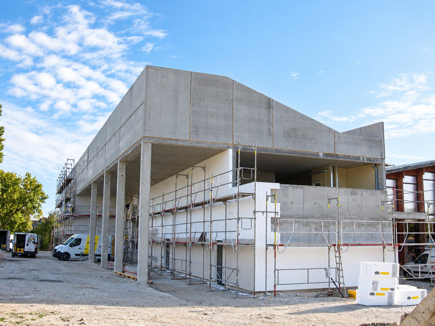 Un bâtiment en béton en construction, dont la façade est équipée d'échafaudages et d'isolant blanc, avec des camionnettes et des blocs d'isolant au premier plan.