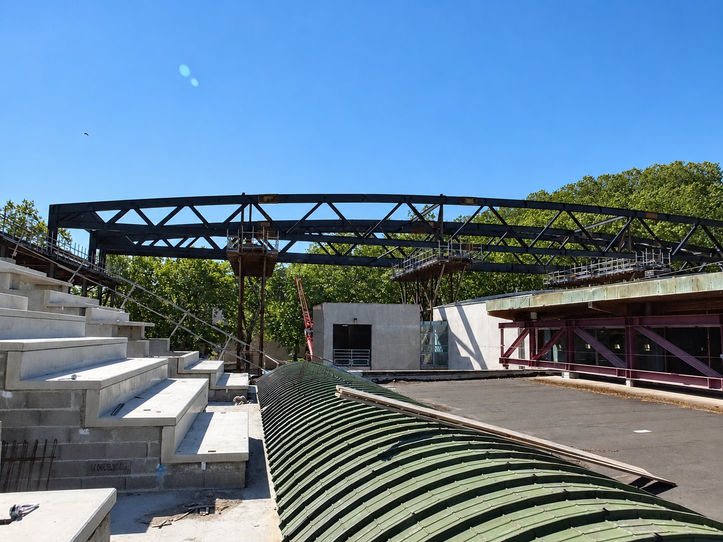 Charpente métallique sombre en construction au-dessus de gradins en béton et d'une toiture voûtée verte, sous un ciel bleu.