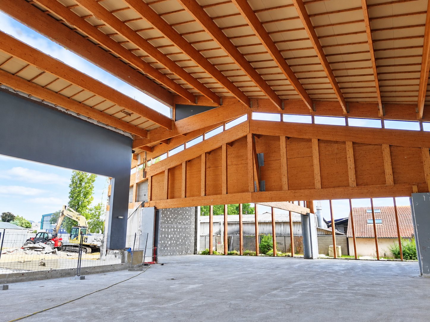 Vue intérieure d'un grand bâtiment en chantier avec une charpente en bois apparente, une dalle en béton et des engins de chantier visibles à l'extérieur.