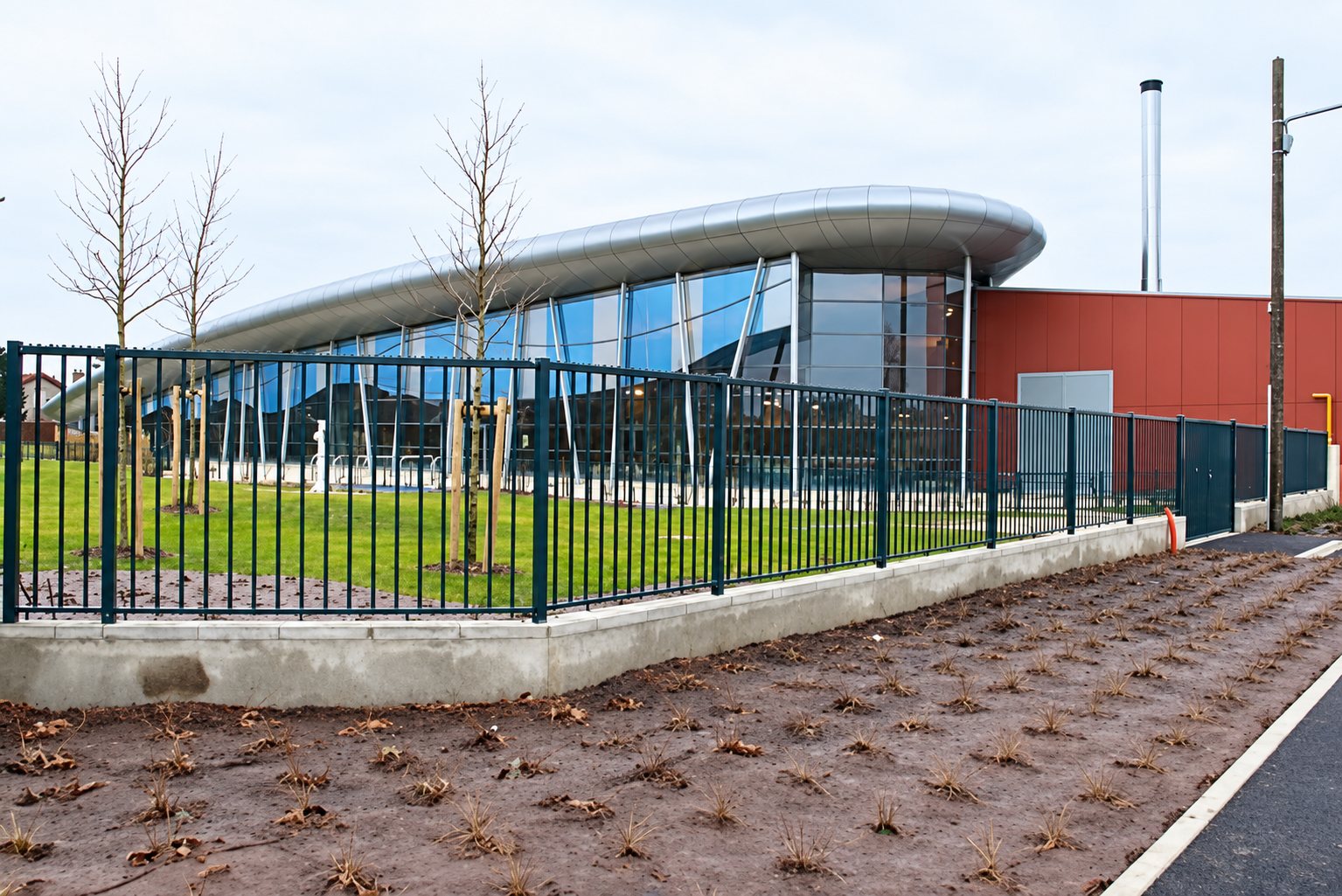 Une clôture métallique noire sur un muret en béton borde une pelouse et un bâtiment moderne à toit courbe et façade vitrée, avec un mur rouge à droite.