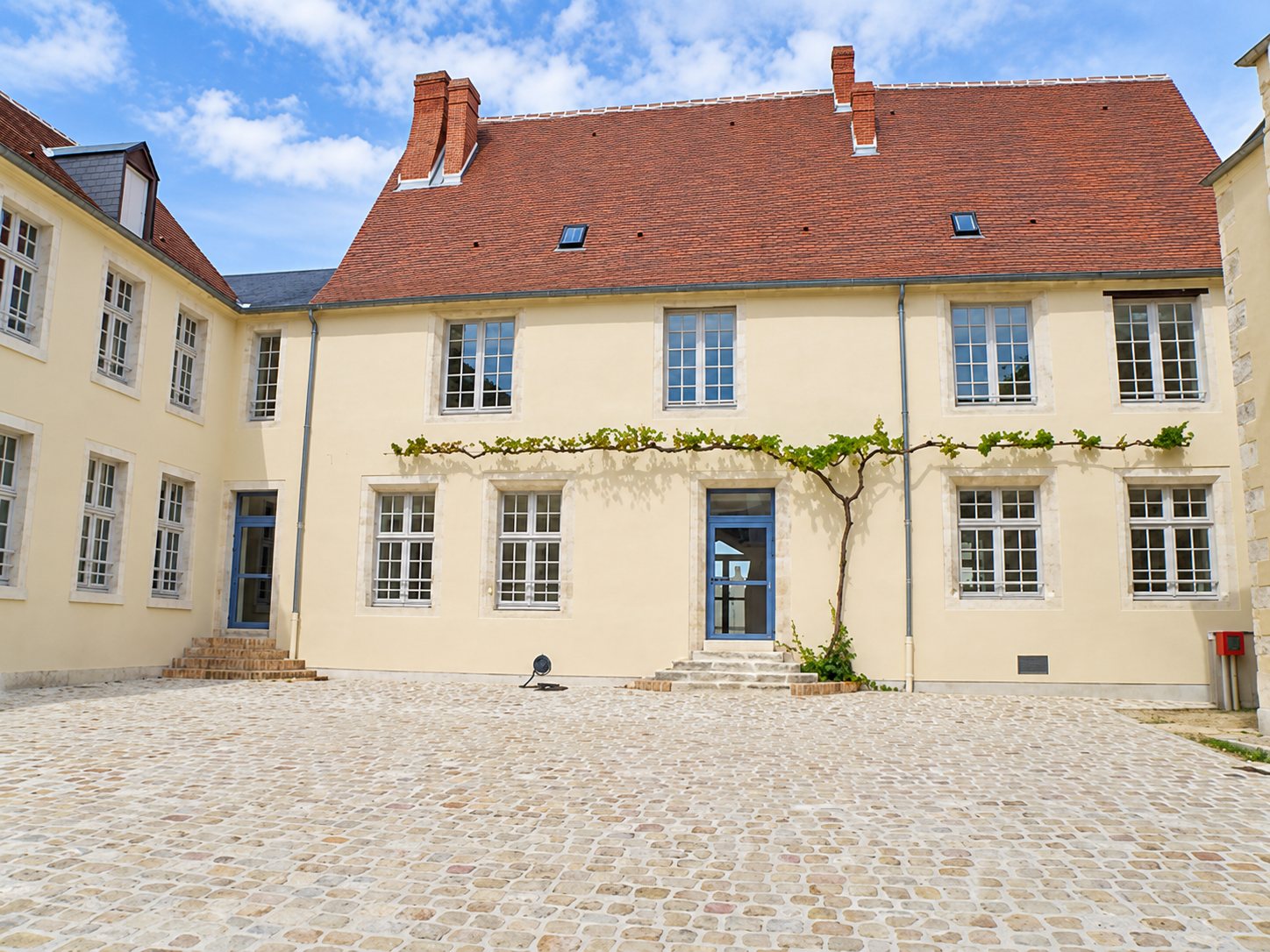 Façade rénovée d'un bâtiment ancien aux murs crème et au toit de tuiles rouges, avec une vigne palissée et une cour pavée au premier plan.