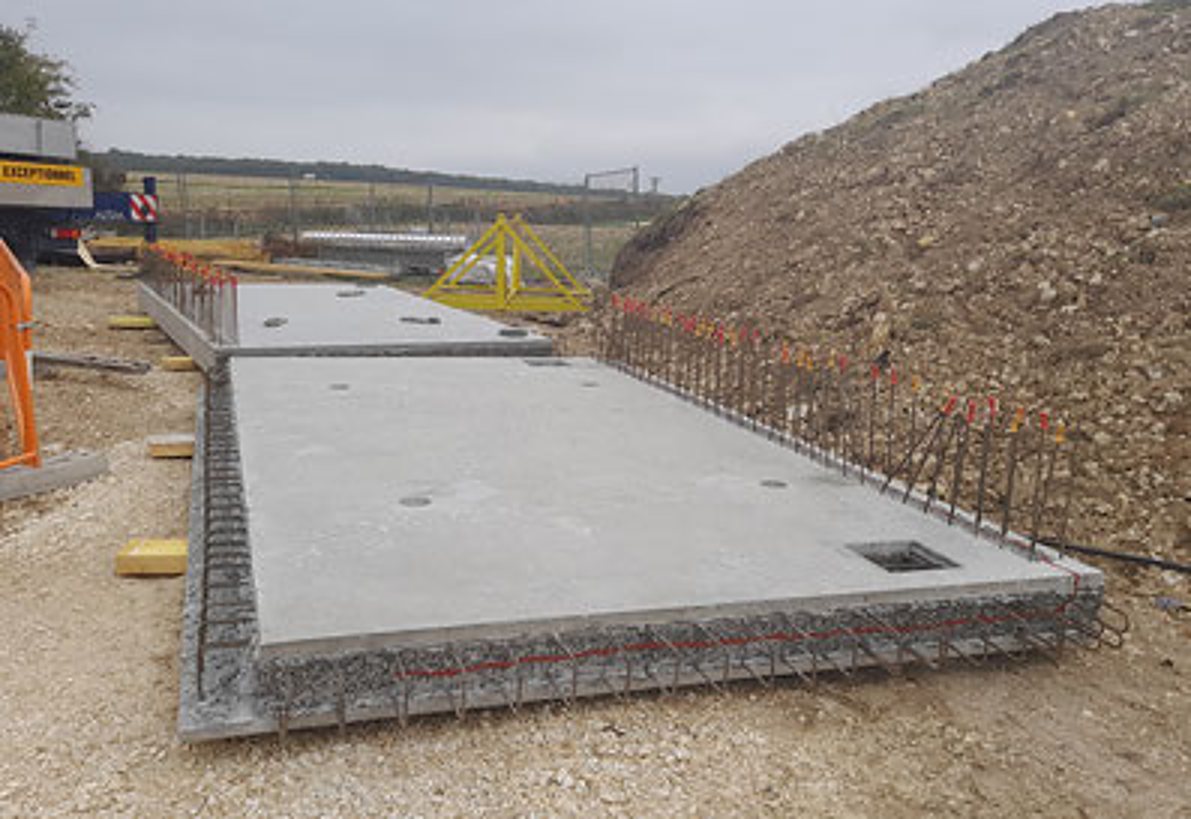 Deux grandes dalles en béton avec armatures apparentes reposent sur un sol de graviers, devant un talus de terre et de cailloux sur un chantier.