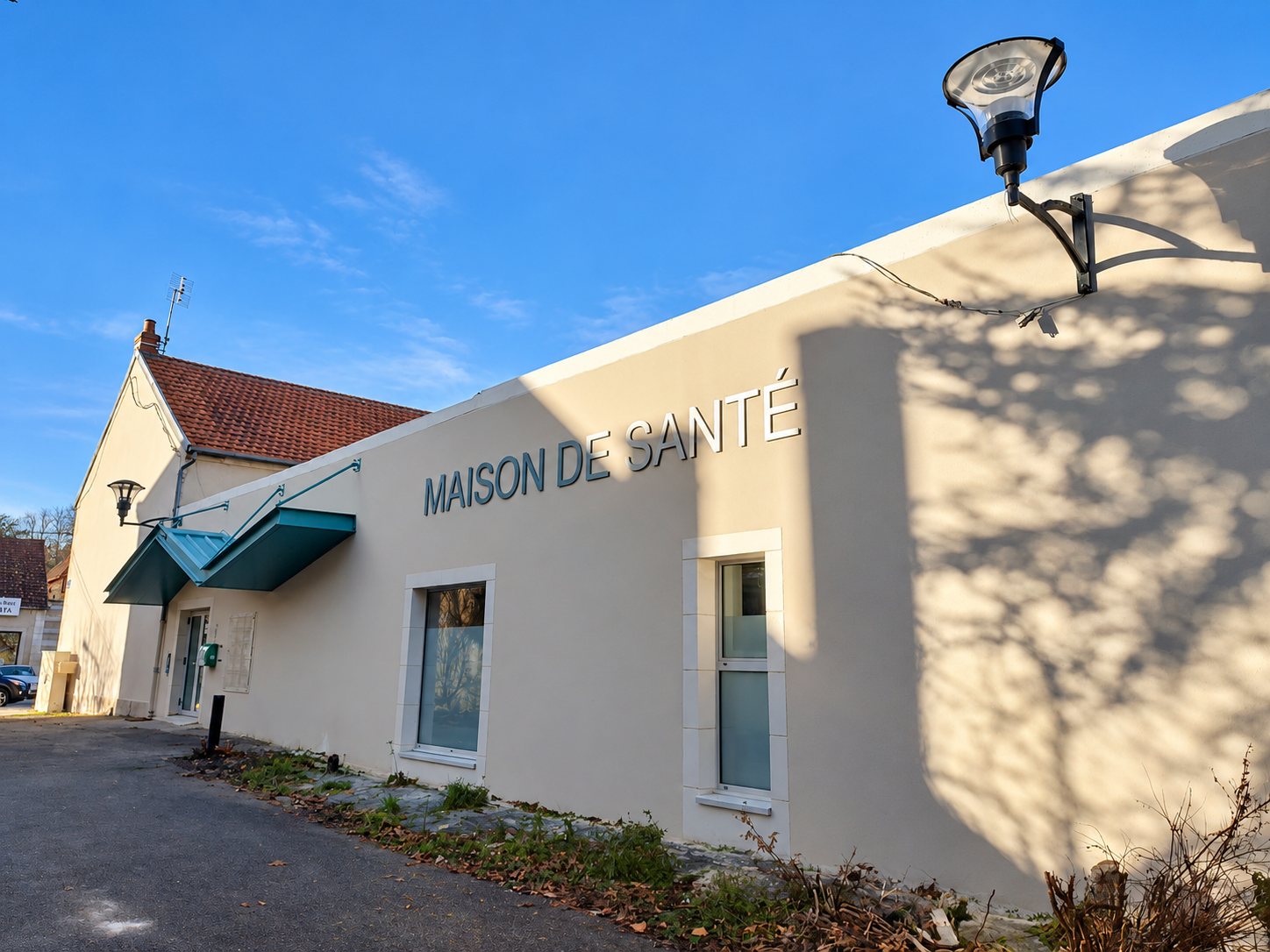 Façade beige d'une maison de santé portant l'enseigne en lettres métalliques, avec un auvent bleu et un lampadaire, sous un ciel bleu.