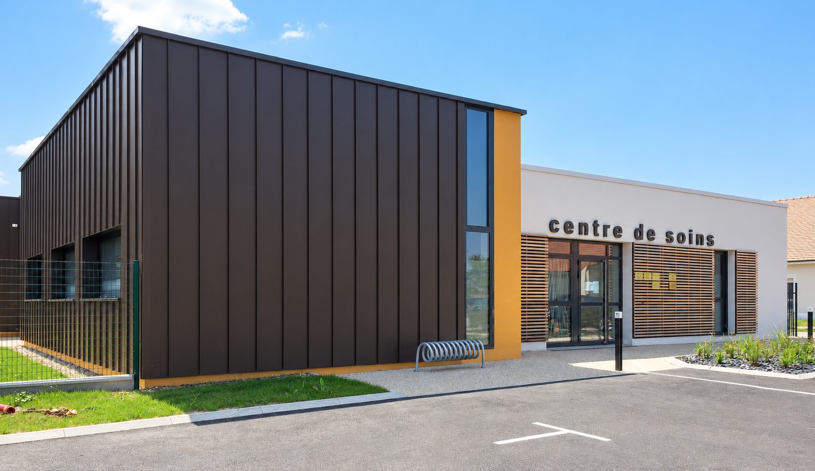 Bâtiment moderne d'un centre de soins avec un bardage métallique brun foncé, une touche orange, des brise-soleil en bois et un parking au premier plan.