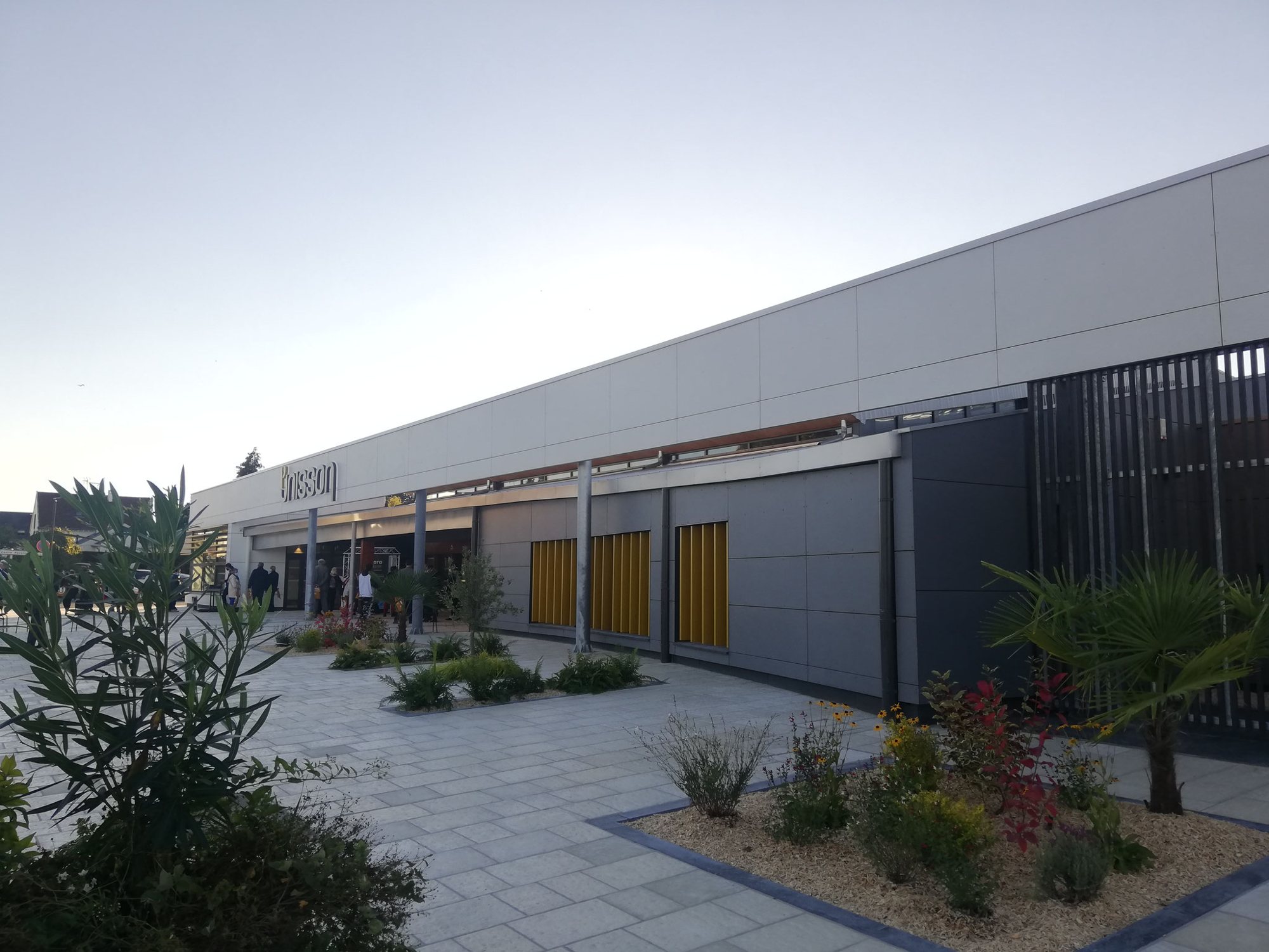 Façade d'un bâtiment commercial moderne à panneaux gris et jaunes, avec un parvis dallé orné de massifs de plantes et de palmiers.