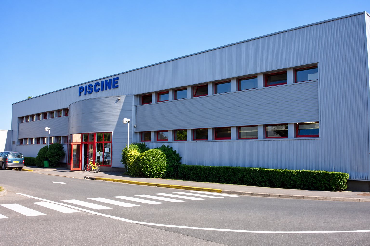 Façade d'un bâtiment de piscine en bardage gris avec l'enseigne bleue PISCINE, une entrée à cadre rouge, des haies et un passage piéton au premier plan.