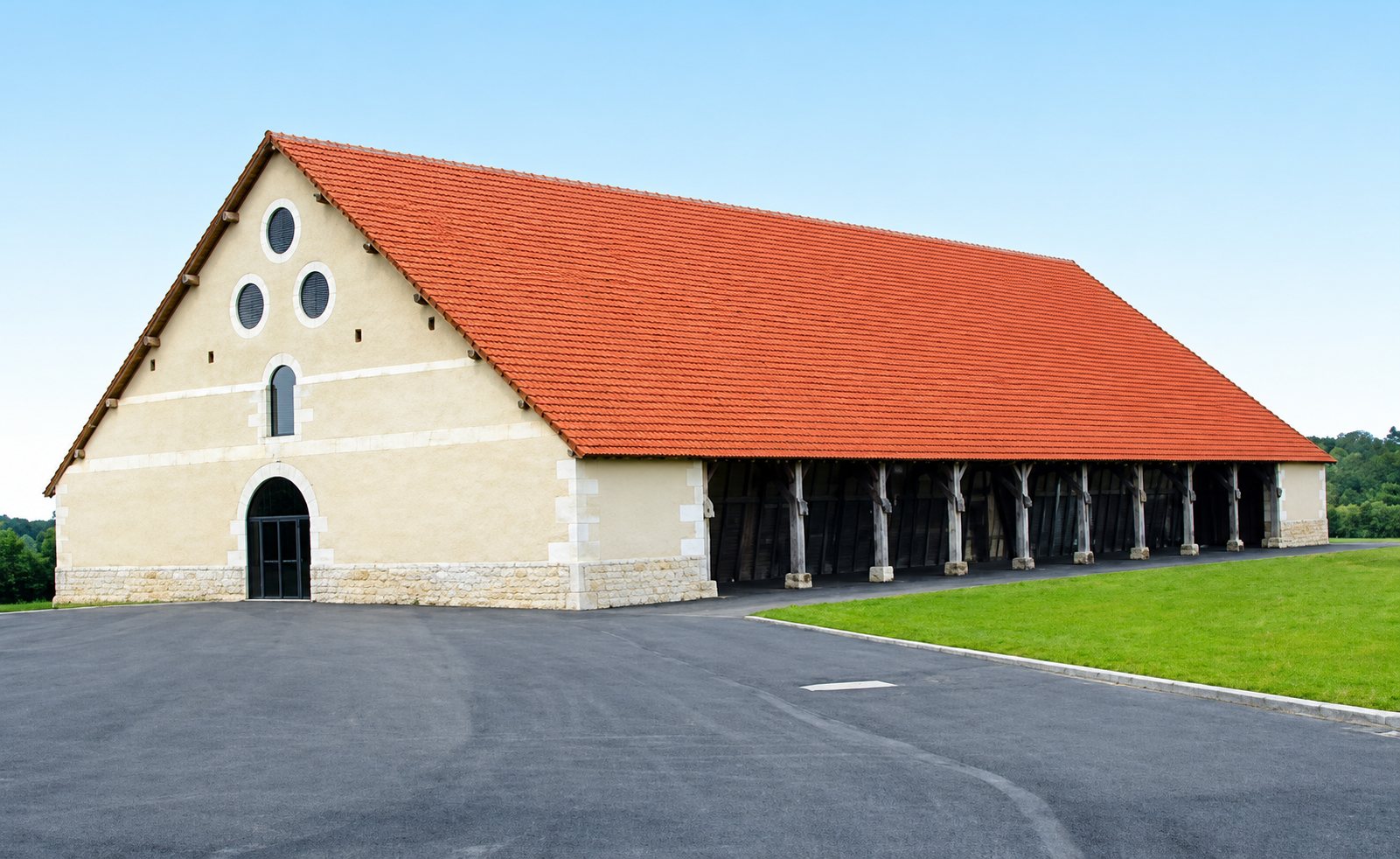 Une grande grange en pierre claire au toit de tuiles orange, ouverte sur un côté par des poteaux en bois, se dresse près d'un parking goudronné et d'une pelouse.