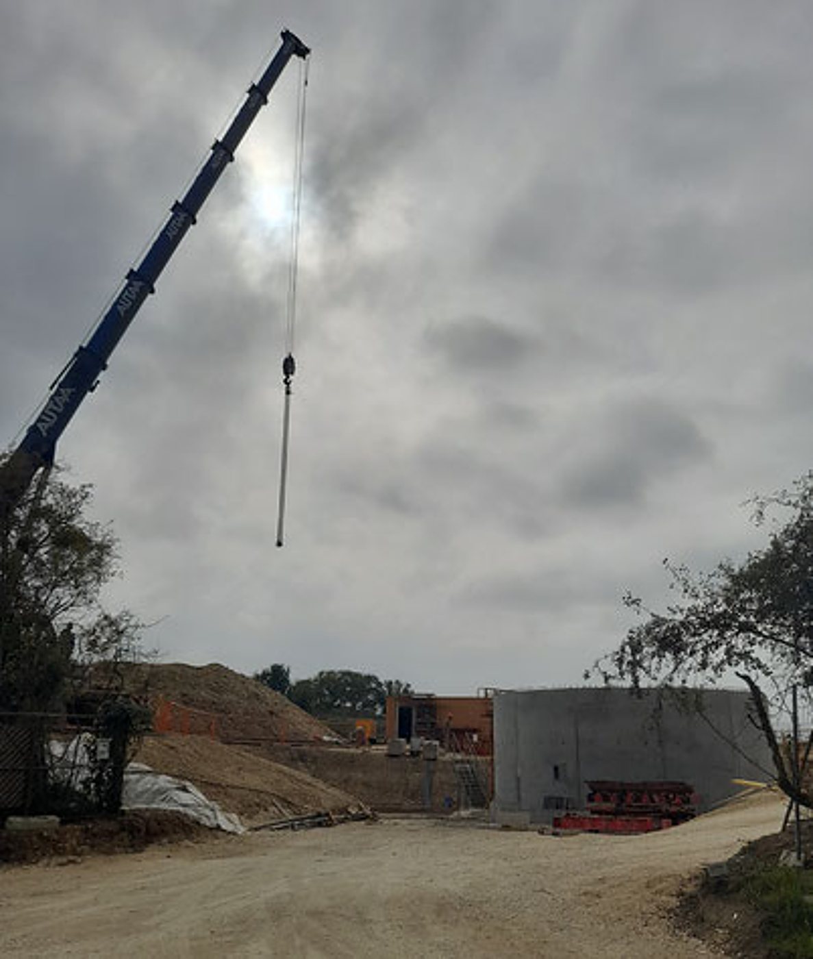 Une grue mobile bleue déploie sa flèche au-dessus d'un chantier avec des tas de terre, un grand mur en béton et un chemin de terre sous un ciel couvert.