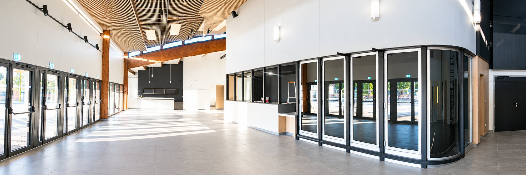 Vue panoramique d'un grand hall moderne et lumineux, avec de larges baies vitrées, des poutres en bois et un plafond acoustique en lames de bois.