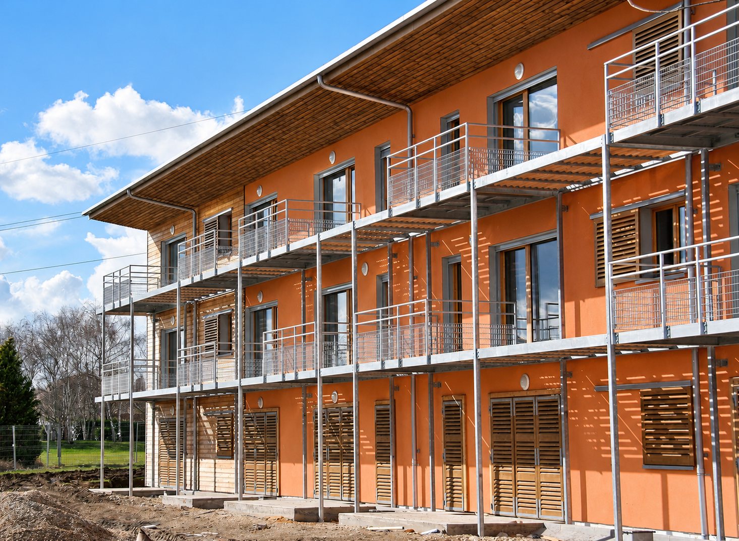 Façade d'un immeuble récent aux murs orangés, équipé de balcons à garde-corps métalliques, de volets en bois et d'une grande toiture débordante, avec un terrain de chantier au premier plan.