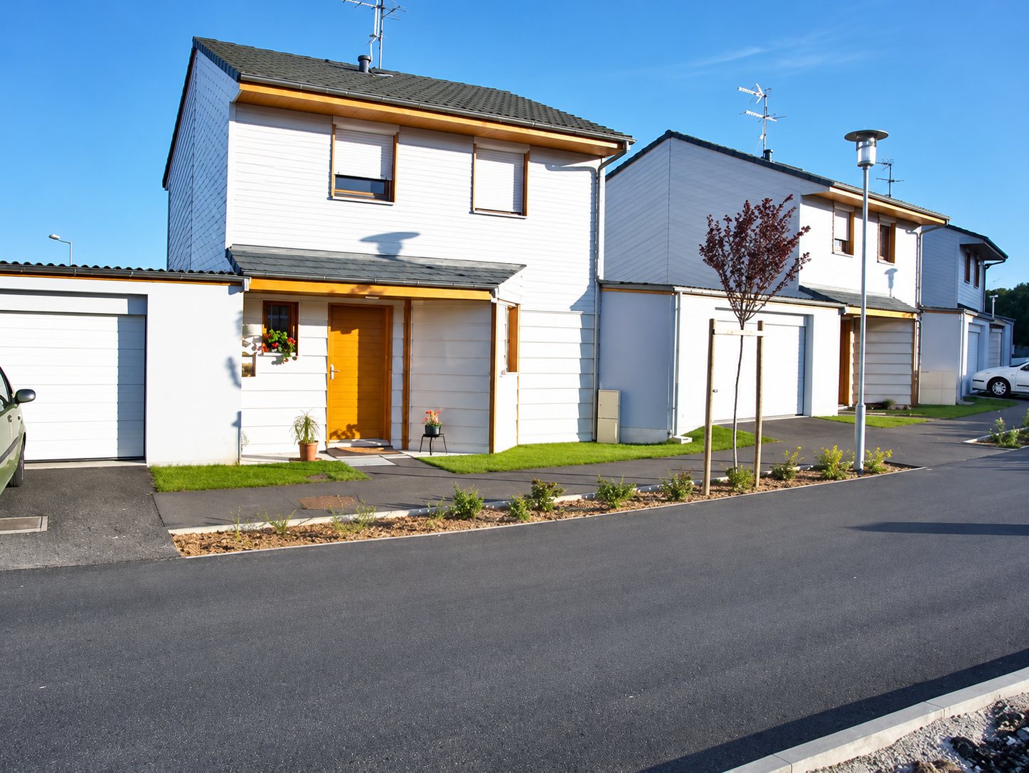 Des maisons neuves aux façades blanches et aux portes orange bordent une route fraîchement goudronnée sous un ciel bleu.