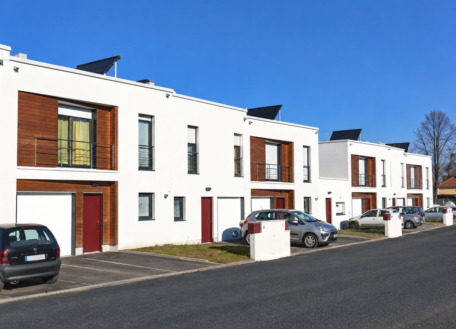 Un ensemble de maisons de ville modernes aux façades blanches et bois, avec des portes rouges, devant un parking où stationnent quelques voitures.