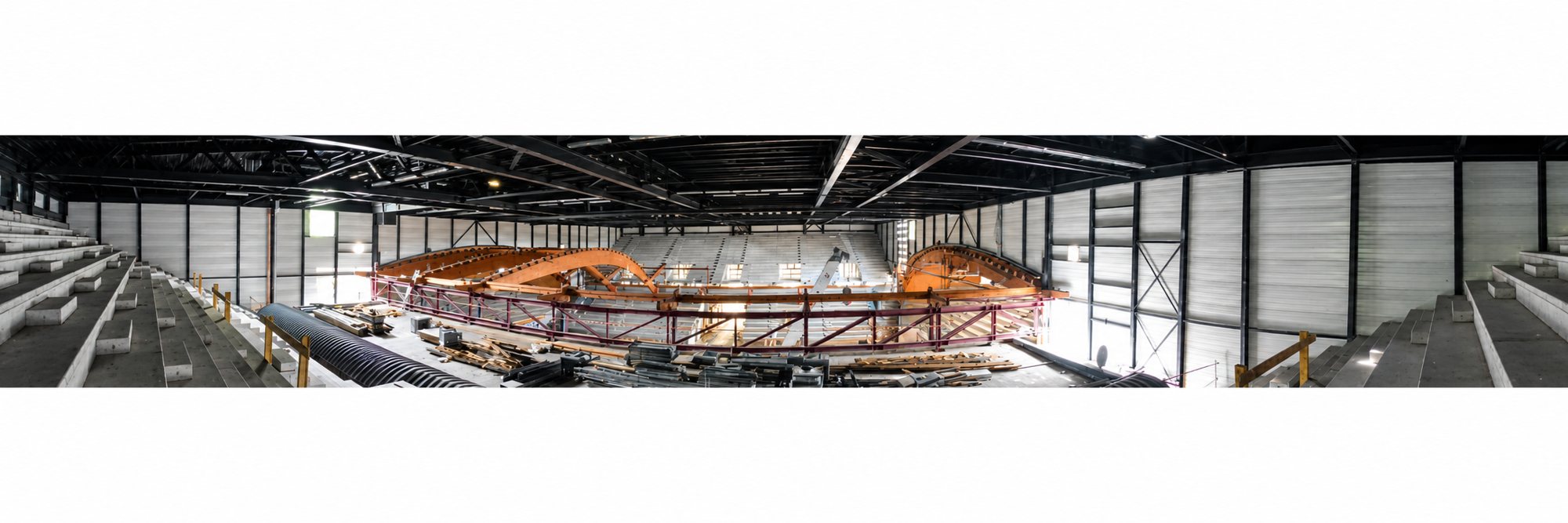 Photo panoramique de l'intérieur d'une grande salle en chantier, avec des arches de charpente en bois, des gradins et des murs en panneaux métalliques.