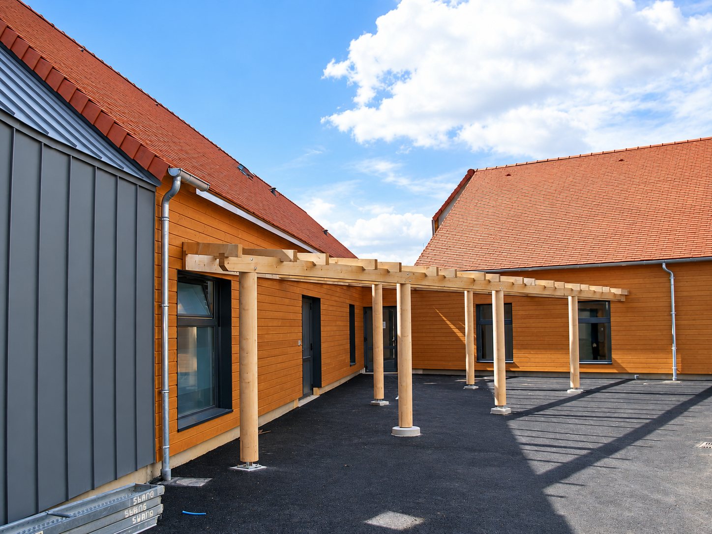 Une longue pergola en bois sur poteaux borde une cour en enrobé noir entre deux bâtiments à bardage bois, toits en tuiles rouges et façade en zinc gris.
