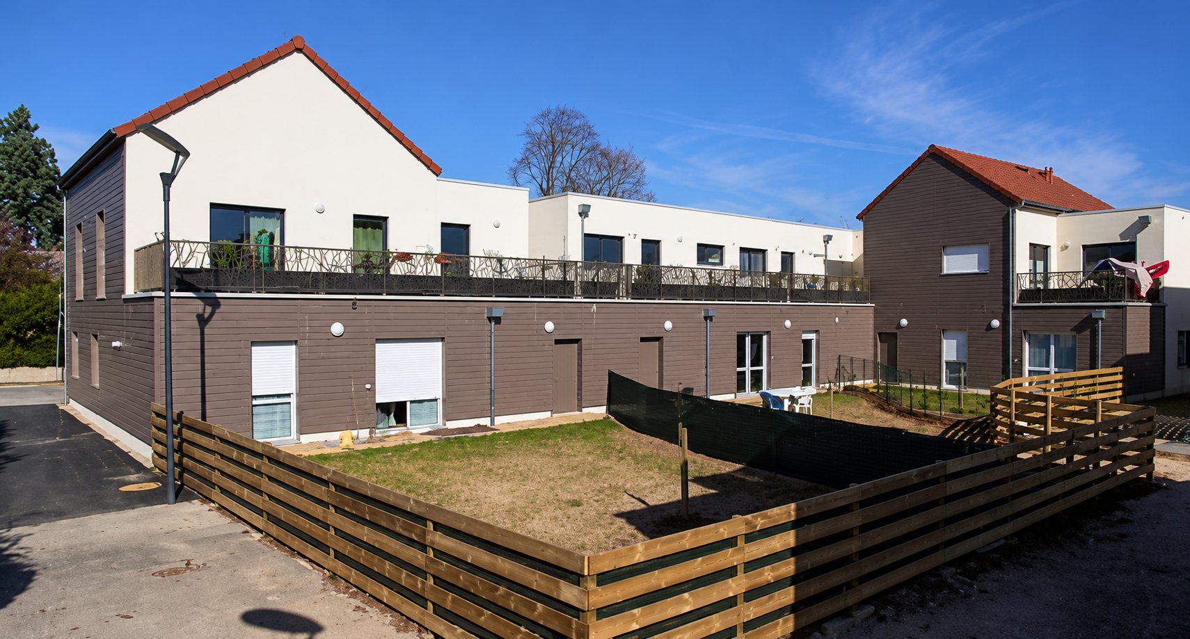 Façade d'une résidence récente aux murs blancs et bruns, avec balcons, terrasses et clôtures en bois autour de petits jardins, sous un ciel bleu.