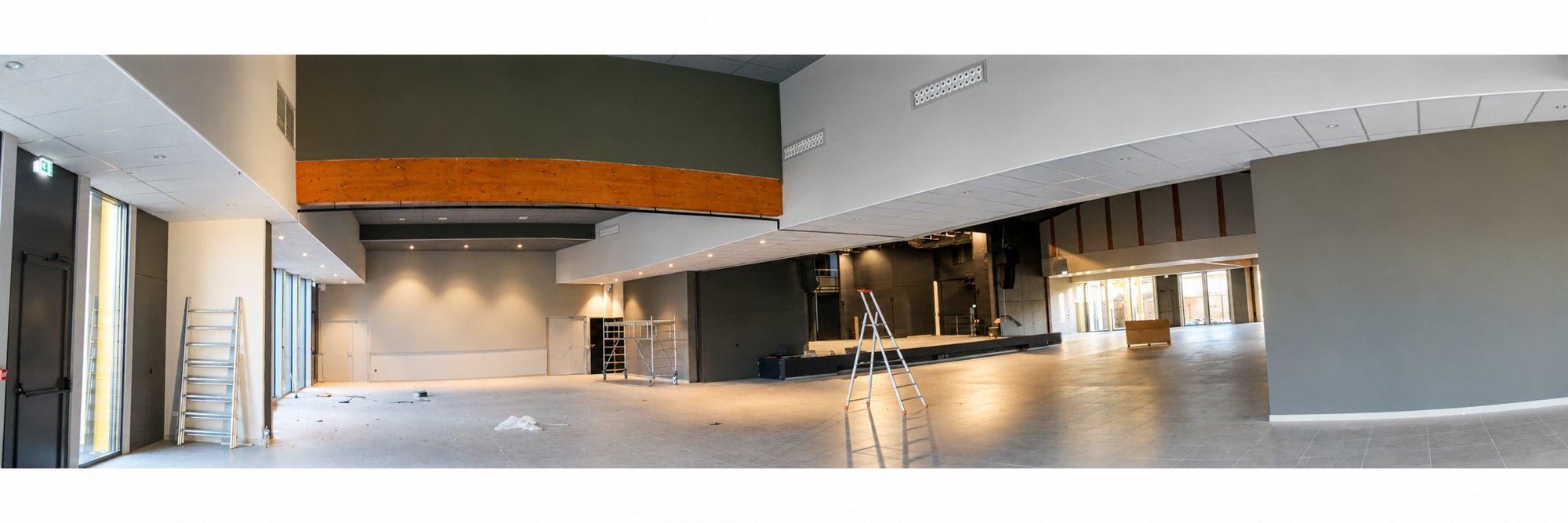 Vue panoramique d'une grande salle presque terminée avec une scène au fond, des échelles et un échafaudage, sans mobilier.