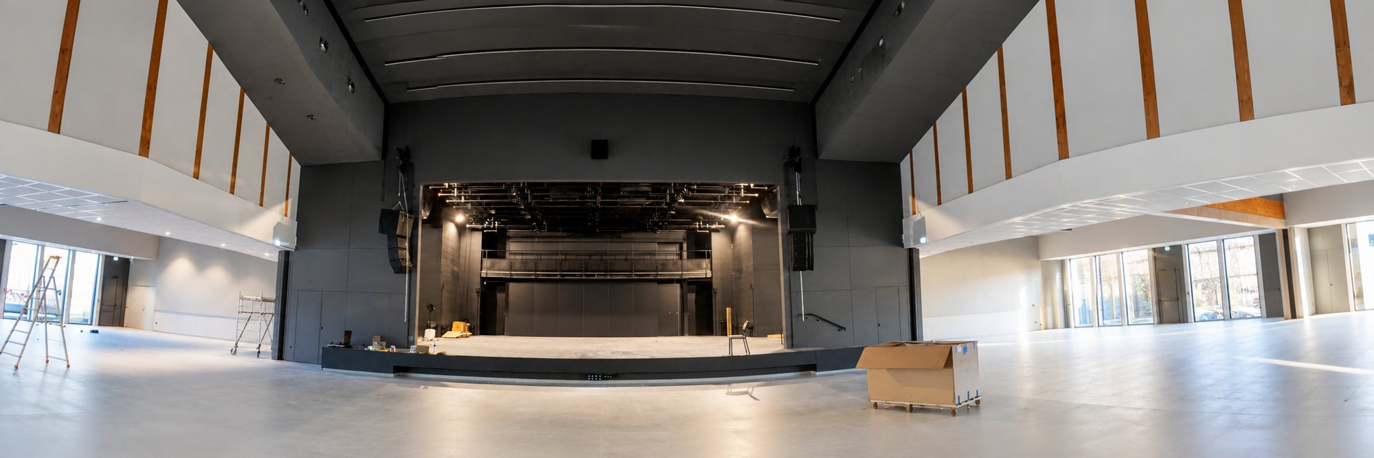Photo panoramique d'une grande salle de spectacle vide, avec une scène au fond, un escabeau et des cartons au sol, lors de travaux de finition.
